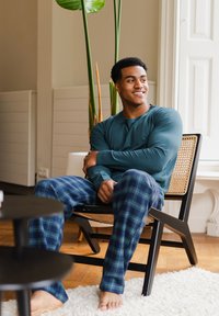 Teal langemouwen shirt en blauwe geruite pyjamabroek gedragen door een persoon die op een houten stoel zit, met een zwarte tafel en groen aanwezig op de achtergrond.