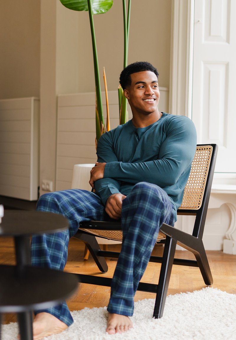 Teal langemouwen shirt en blauwe geruite pyjamabroek gedragen door een persoon die op een houten stoel zit, met een zwarte tafel en groen aanwezig op de achtergrond.