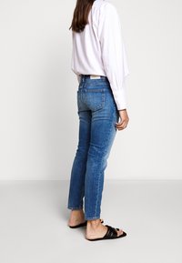 Jean bleu taille haute à la coupe ajustée avec une finition légèrement délavée. Associé à une chemise blanche à manches longues et des sandales noires à enfiler.