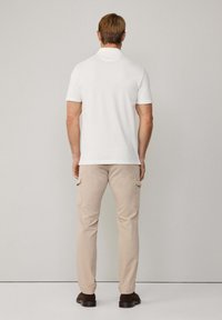 Polo bianco, maniche corte, design semplice. Pantaloni cargo beige con tasche laterali. Scarpe marroni. Sfondo uniforme.