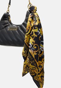 Černá prošívaná kabelka se zlatou kovovou výbavou a vzorovaným černým a zlatým šátkem přivázaným k uchu. Na přední straně je umístěno logo značky.