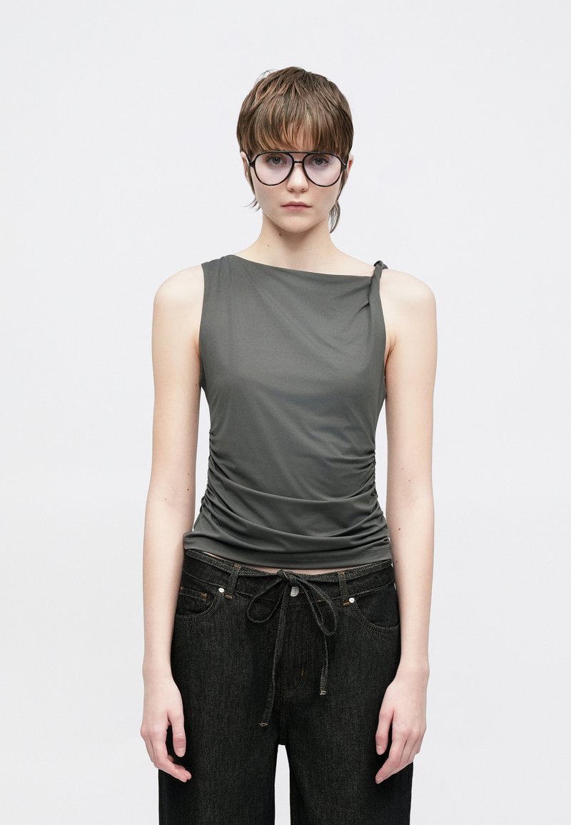 Haut gris sans manches en tissu lisse et extensible, avec des fronces sur le côté et un large décolleté. Associé à un jean foncé.