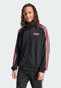 Veste zippée noire avec des rayures rouges et grises sur les manches, un patch logo Adidas et des poignets côtelés, fabriquée à partir d'un matériau léger.