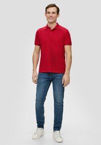 Rotes Polohemd mit kurzen Ärmeln und Kragen, kombiniert mit blauen Jeans und weißen Sneakers. Weicher Baumwollstoff mit taillierter Passform.