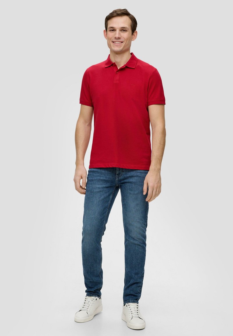 Rotes Polohemd mit kurzen Ärmeln und Kragen, kombiniert mit blauen Jeans und weißen Sneakers. Weicher Baumwollstoff mit taillierter Passform.