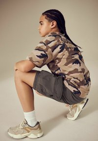 Chemise à manches courtes avec motif camouflage dans des tons beiges et verts, associée à un short gris et des baskets crème avec des accents en tan.