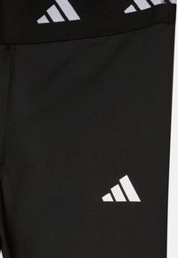 Leggings deportivos negros con una textura suave, que presentan un logo blanco de Adidas. La cintura incluye un patrón gráfico en blanco y negro.