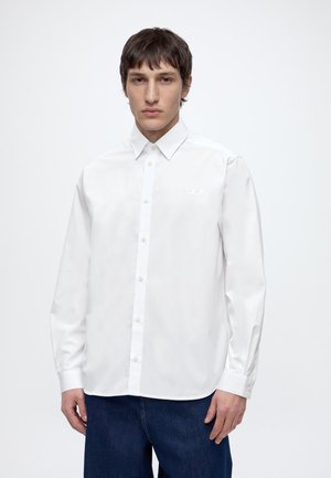 Giovane uomo con capelli castani corti che indossa una camicia bianca abbottonata e pantaloni blu scuro, in piedi davanti a uno sfondo chiaro e uniforme.