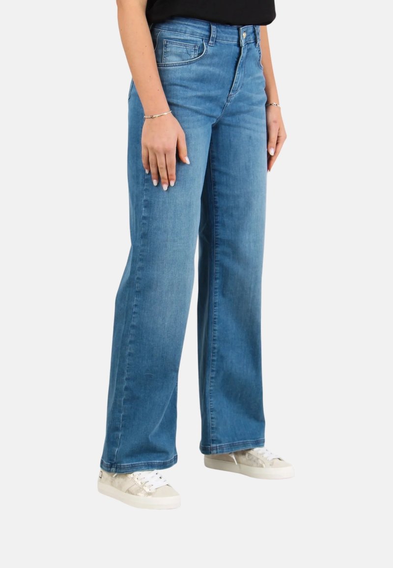 Jeans a gamba larga in denim azzurro chiaro con vita media, dotati di tasche frontali e una texture liscia. Indossati con sneakers bianche.