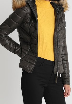 Personne portant une veste matelassée noire avec capuche bordée de fourrure, un pull à col roulé côtelé jaune et un pantalon noir.