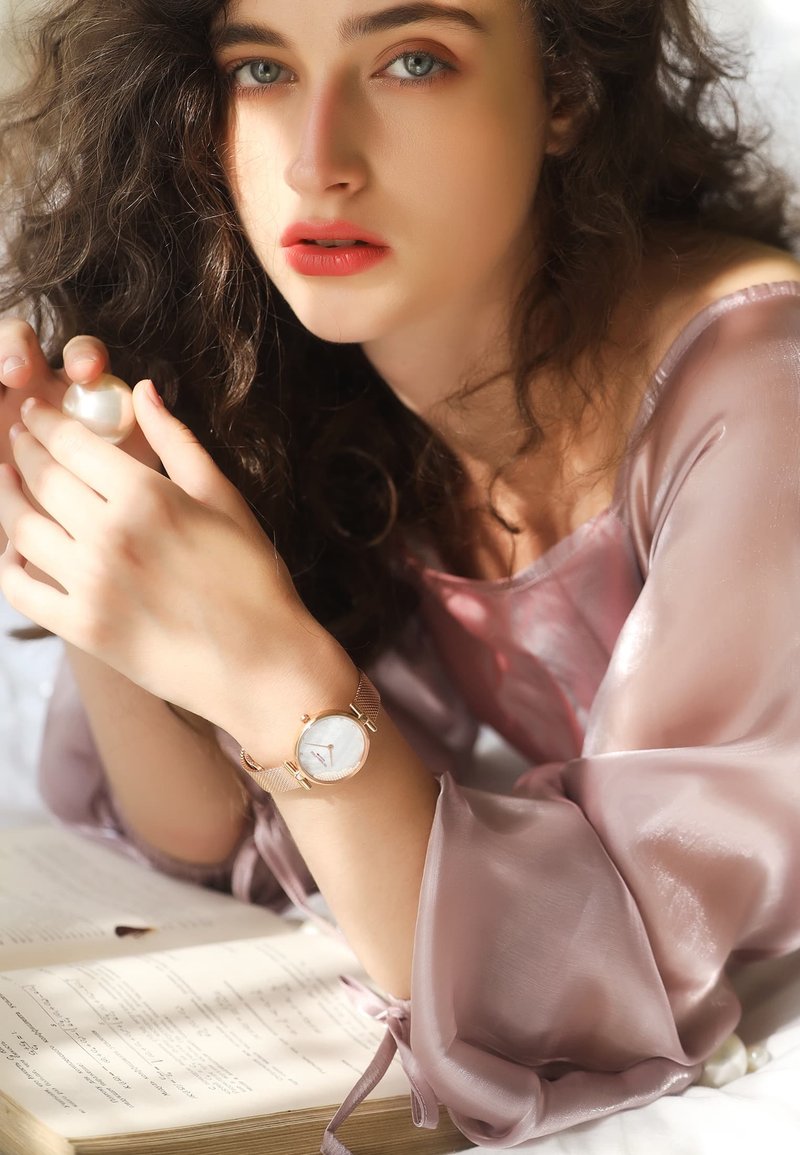 Jeune femme aux cheveux bouclés portant un chemisier transparent lavande, tenant une perle, portant une montre en or rose, reposant sur un livre ouvert.