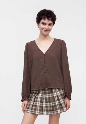 Junge Frau mit kurzen lockigen Haaren trägt eine braune Bluse mit langen Ärmeln und einen plissierten braun-weißen karierten Minirock, steht vor einem einfarbigen Hintergrund.