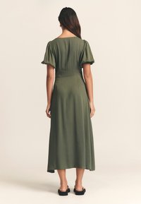 Robe vert olive, longueur genou, avec des manches bouffantes et une couture à la taille. Tissu lisse avec une texture douce. Le dos présente un décolleté large.