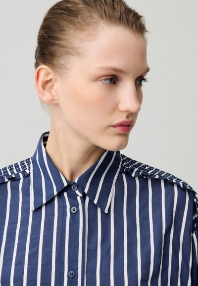 Camisa a rayas azul marino y blanca con cuello abotonado, que cuenta con un detalle texturizado en los hombros y sutiles volantes a lo largo de los hombros.