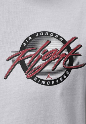 Logo Air Jordan Flight avec script rouge sur cercle noir et rayures, texte "Depuis 1984" sur fond de tissu blanc.