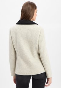 Eine Frau trägt eine hellgraue Wolljacke mit schwarzem Kragen, die von hinten zu sehen ist und vor einem schlichten weißen Hintergrund steht.