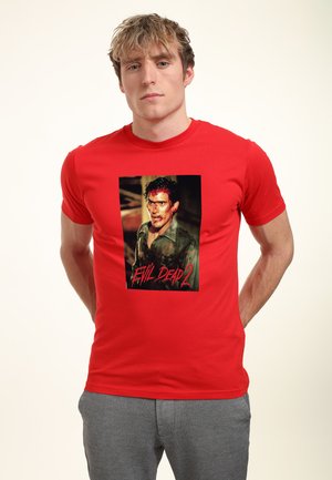 Rød bomulds T-shirt med et stort grafisk motiv af en karakter med mørkt hår og en lys skjorte, med "Evil Dead 2" tekst i rødt under billedet.