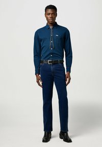 Dark blue button-up shirt with a pointed collar and chest pocket, paired with dark jeans and black ankle boots. Bolo tie accent.