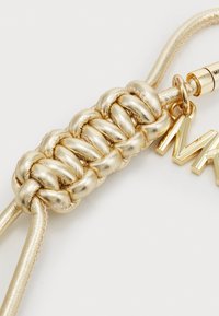 Corde tressé en or avec un éclat métallique, comportant un charme doré sur lequel est inscrit "MK". Texture lisse avec un design élégant.