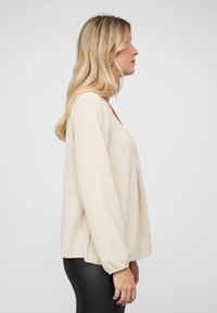 Beige langslavenshirt met een gestructureerde afwerking, relaxte pasvorm, ronde halslijn en elastische manchetten, gecombineerd met aansluitende zwarte leggings.