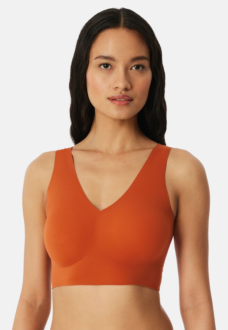 Femme aux longs cheveux foncés portant un brassière de sport orange sans manches avec un décolleté en V, face à l'avant sur un fond blanc uni.