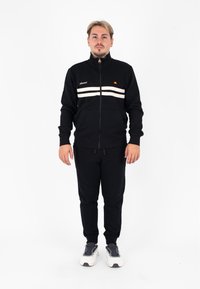Tuta nera realizzata in tessuto morbido, con giacca con zip e strisce bianche e logo, abbinata a pantaloni coordinati. Modello in piedi davanti.