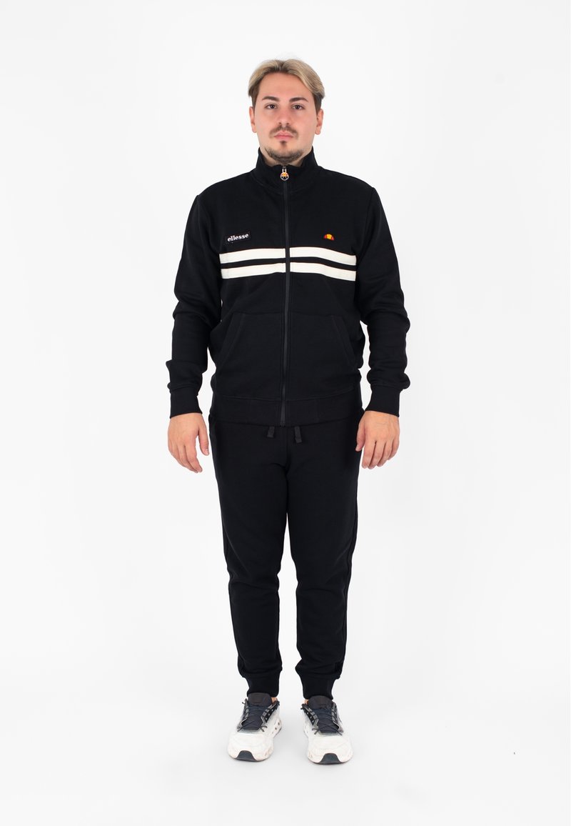 Tuta nera realizzata in tessuto morbido, con giacca con zip e strisce bianche e logo, abbinata a pantaloni coordinati. Modello in piedi davanti.