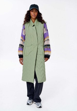 Jasnozielona pikowana kamizelka z załamaną kołnierzem i przednimi kieszeniami, nałożona na wielokolorowy sweter z dzianiny. Ciemne spodnie i trampki dopełniają stylizację.