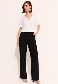 Biała bluzka z dekoltem w serek i krótkimi rękawami w połączeniu z czarnymi spodniami o wysokim stanie; model noszący beżowe sandały i złote kolczyki w kształcie kółek jako akcenty.