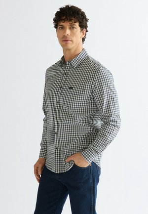 Mann mit lockigem Haar trägt ein blau-weiß kariertes Hemd mit Knöpfen und dunkelblaue Jeans und steht mit einer Hand in der Tasche.