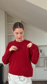 Red cable-knit sweater with a ribbed neckline and cuffs, featuring a small logo detail on the chest. The texture is thick and cozy.
