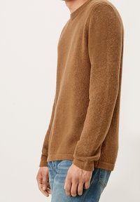 Hnědý pletený svetr s kulatým výstřihem, texturovaná látka a dlouhými rukávy, v kombinaci s světle modrými džínami. Boční pohled ukazuje manžety rukávů a lem.