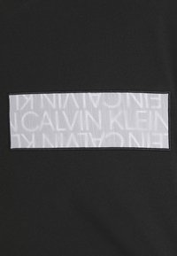 Černá mikina s kulatým výstřihem, která má obdélníkovou bílou nášivku s textem "CALVIN KLEIN" v opakovaném vzoru, se šitými okraji a hladkým povrchem.