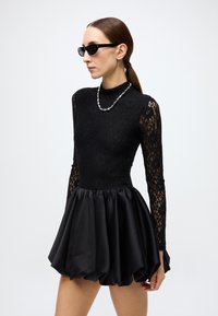 Black dress featuring a lace bodice and flared satin skirt. Long lace sleeves; styled with black sunglasses and a silver bead necklace.
