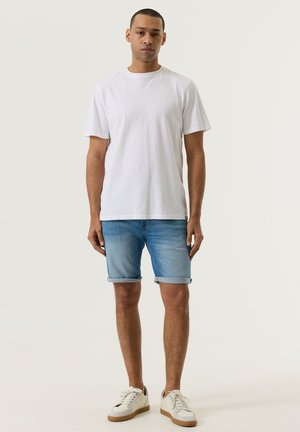 Man die staat, gekleed in een effen wit t-shirt, opgerolde blauwe denim shorts en witte sneakers, tegen een effen achtergrond.
