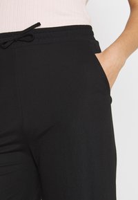 Pantalons noirs à taille élastique avec un détail à cordon, présentant une texture lisse et des poches latérales, présentés sur un fond clair.