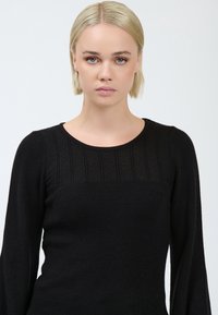 Giovane donna con capelli biondi corti indossa un top nero lavorato a maglia con yoke testurizzato, rivolta verso l'obbiettivo su uno sfondo grigio chiaro uniforme.