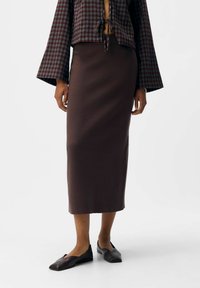 Vrouw in een bruine midi-rok, een donkerbruine geruite blouse met lange mouwen en strikdetail, en donkerbruine platte schoenen die tegen een witte achtergrond staat.