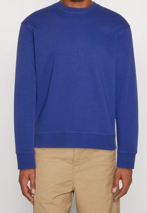 Sweatshirt bleu à col rond en tissu texturé, doté de manches longues et d'un ourlet droit. Porté avec un pantalon beige.