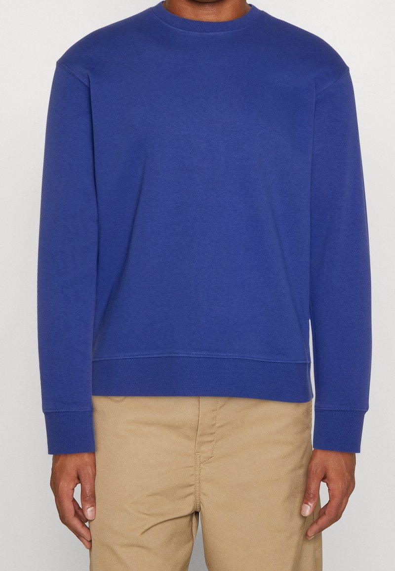 Felpa blu a girocollo realizzata in tessuto texturizzato, con maniche lunghe e orlo dritto. Indossata con pantaloni khaki.