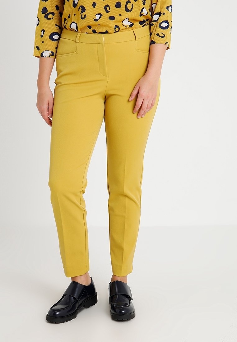 Personne portant un pantalon jaune moutarde, un haut jaune à taches noires et blanches et des mocassins noirs, debout devant un fond clair et uni.