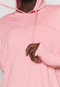 Sweat à capuche rose clair en tissu doux, doté d'une poche kangourou, de poignets en côte et d'un cordon de serrage pour un ajustement optimal.