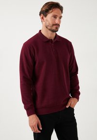 Bordeaux langärmliges Poloshirt mit Kragen, drei Knöpfen, gerippten Bündchen und Saum; aus strukturiertem Stoff. Es werden schwarze Hosen getragen.