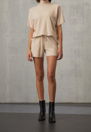 Kvinde iført beige kortærmet top og shorts med snøre, kombineret med sorte ankelstøvler, står op ad en grå tekstureret væg.