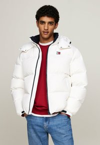 Giacca puffa bianca con chiusura a zip, dotata di cappuccio blu navy, cuciture a contrasto e un piccolo logo sul petto. Indossata sopra un maglione rosso.