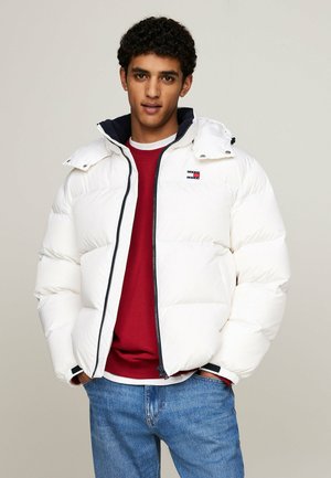 Giacca puffa bianca con chiusura a zip, dotata di cappuccio blu navy, cuciture a contrasto e un piccolo logo sul petto. Indossata sopra un maglione rosso.