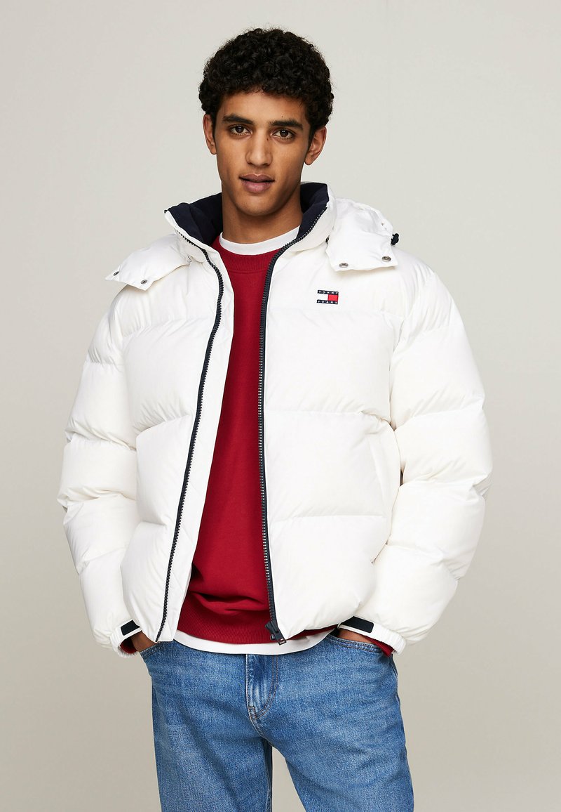 Giacca puffa bianca con chiusura a zip, dotata di cappuccio blu navy, cuciture a contrasto e un piccolo logo sul petto. Indossata sopra un maglione rosso.