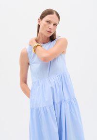 Donna con abito azzurro chiaro senza maniche a balze e bracciale d'oro al polso, posando su uno sfondo bianco uniforme.