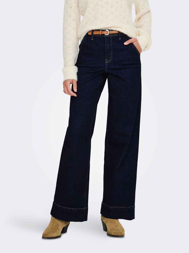 Jean bleu foncé à jambes larges et taille haute, avec des coutures dorées et des poches latérales, associé à un pull en maille texturée couleur crème et des bottes beiges.