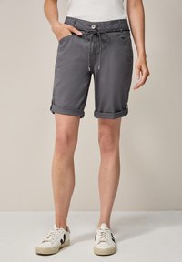 Pantalones cortos grises, hasta la rodilla con puños doblados, con cintura ajustable y bolsillos laterales. Llevados con zapatillas blancas.